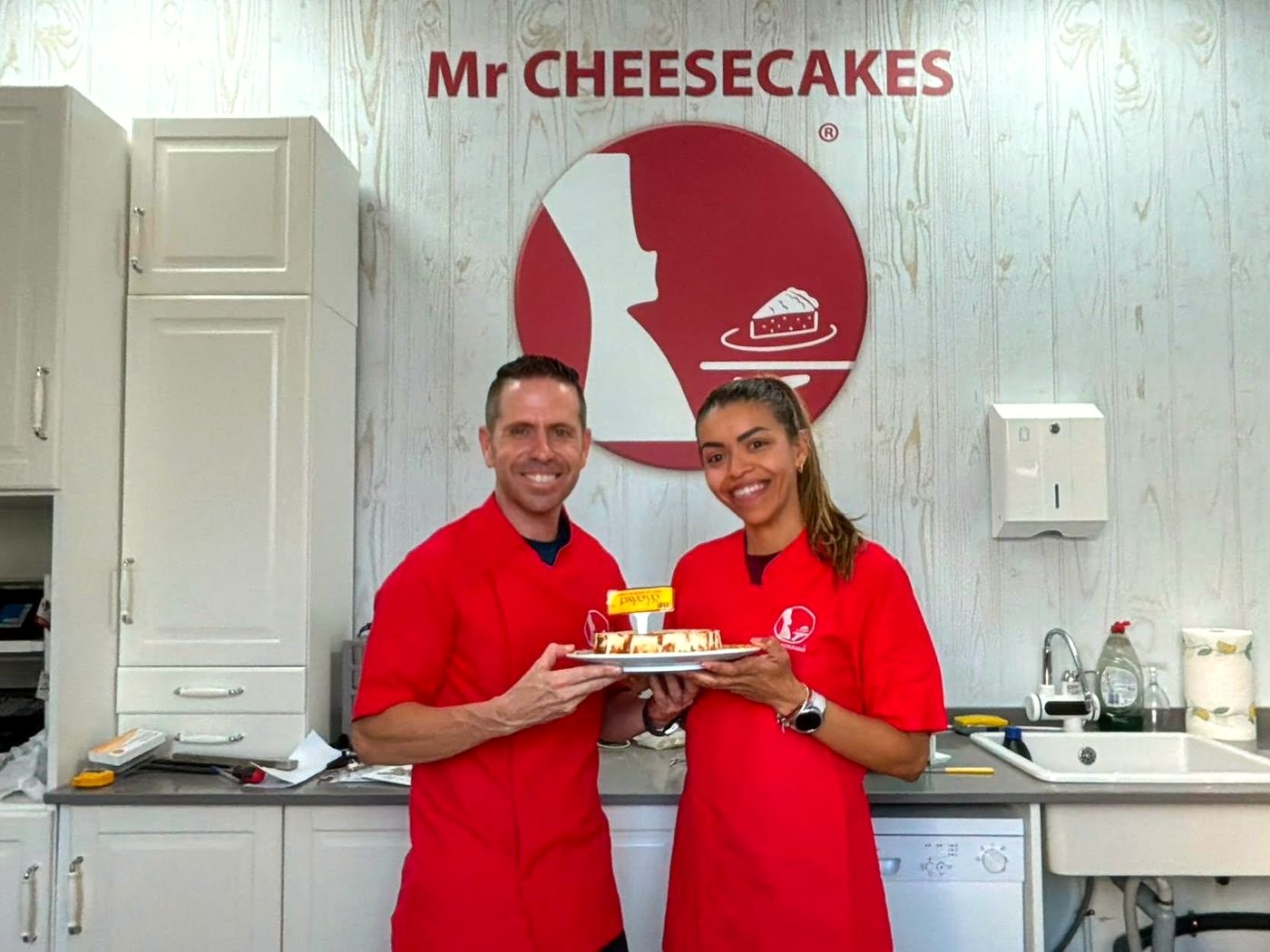 Los dos responsables de Mr Cheesecakes Sanlúcar sonriendo con una tarta de Payoyo en el obrador, bajo el logo de la marca