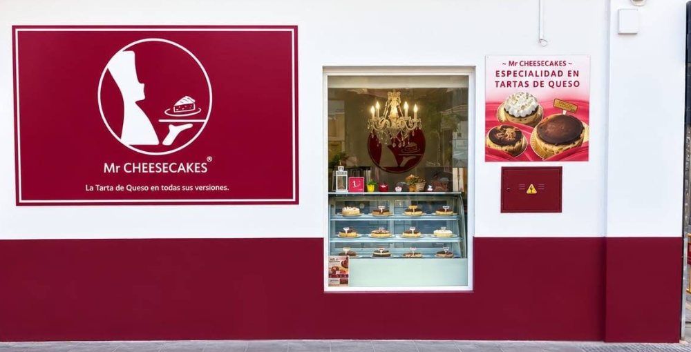 Fachada blanca con zócalo granate de Mr Cheesecakes, con el rótulo y el escaparate lleno de tartas